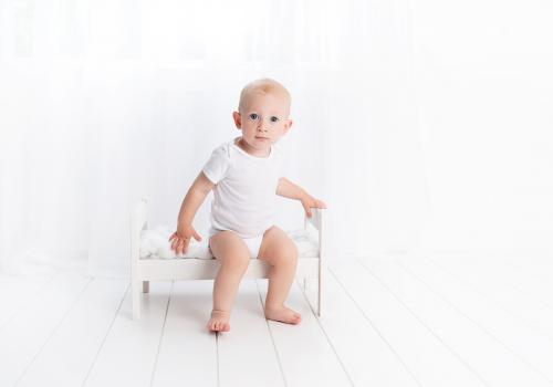 Bebê sentado em caminha branca, com olhar penetrante, vestindo camiseta branca, olha para o centro da imagem