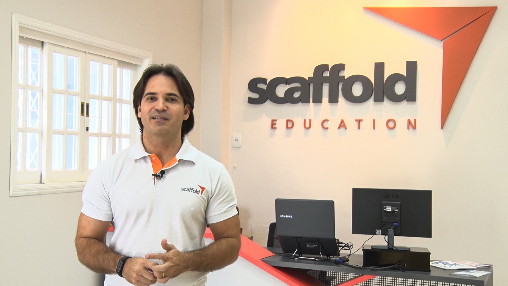 Representante da startup Scaffold Education está de pé, ao lado do logotipo da empresa, que está aplicado em uma parede
