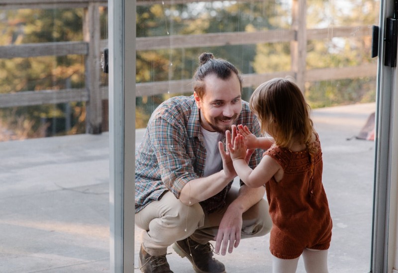 Imagem mostra pai e filha separados por uma porta de vidro ambos com as mãos estendidas